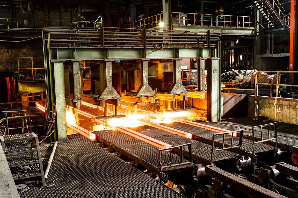 Glowing red-hot steel billets moving through a continuous casting line on roller tables inside a metal processing plant