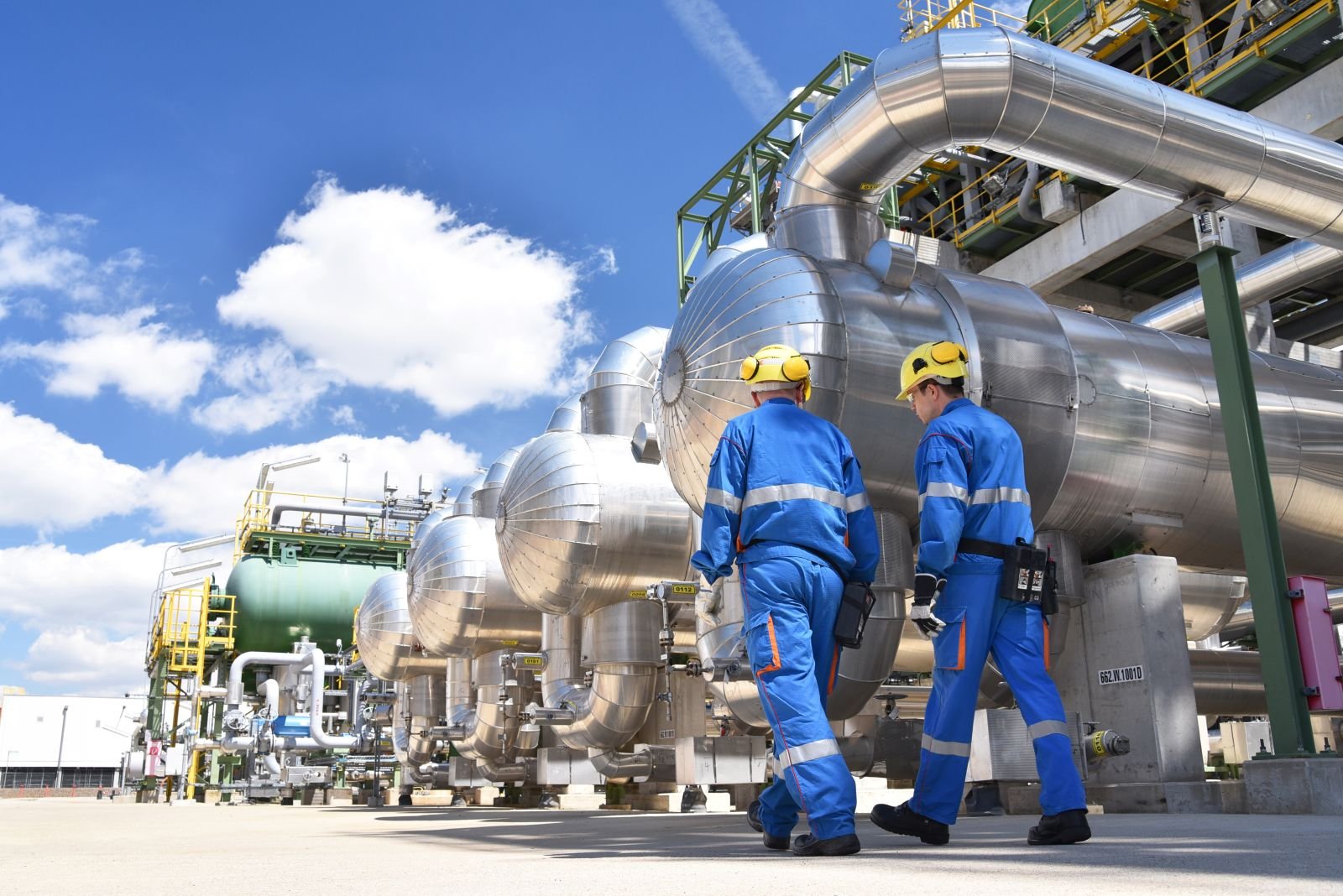 Two workers in safety gear walking past pressure vessels and pipework at a crude oil processing plant
